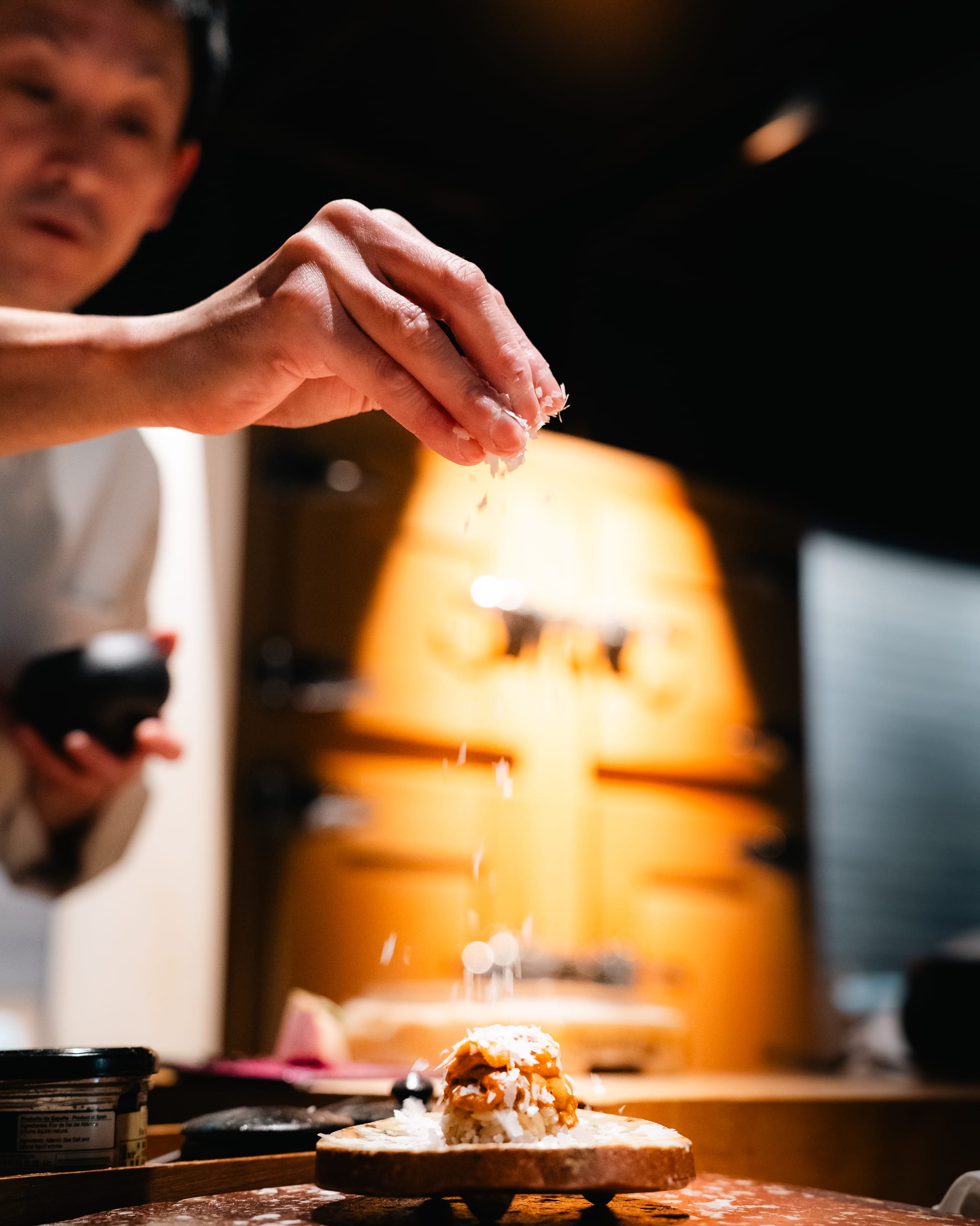 Chef seasoning a dish under warm light