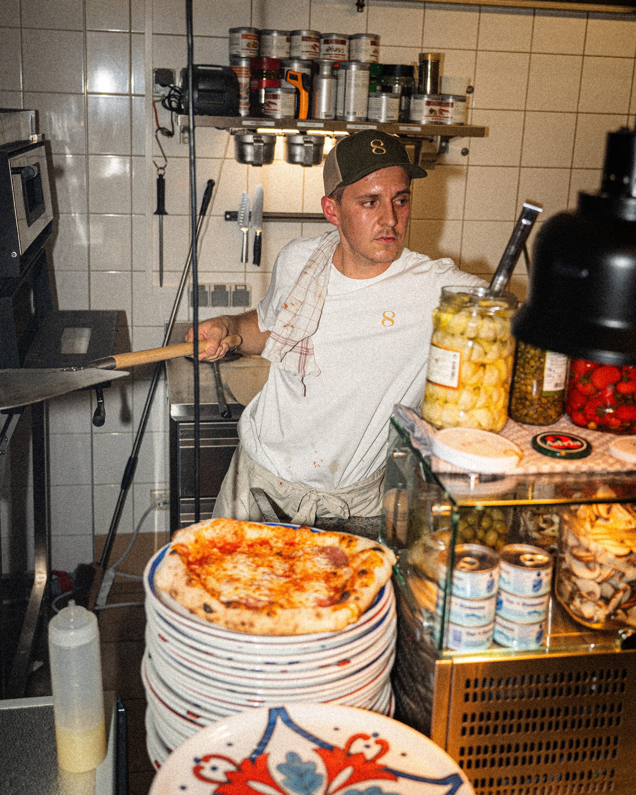 Pizzaiolo beside stacked plates and pizzas in the kitchen